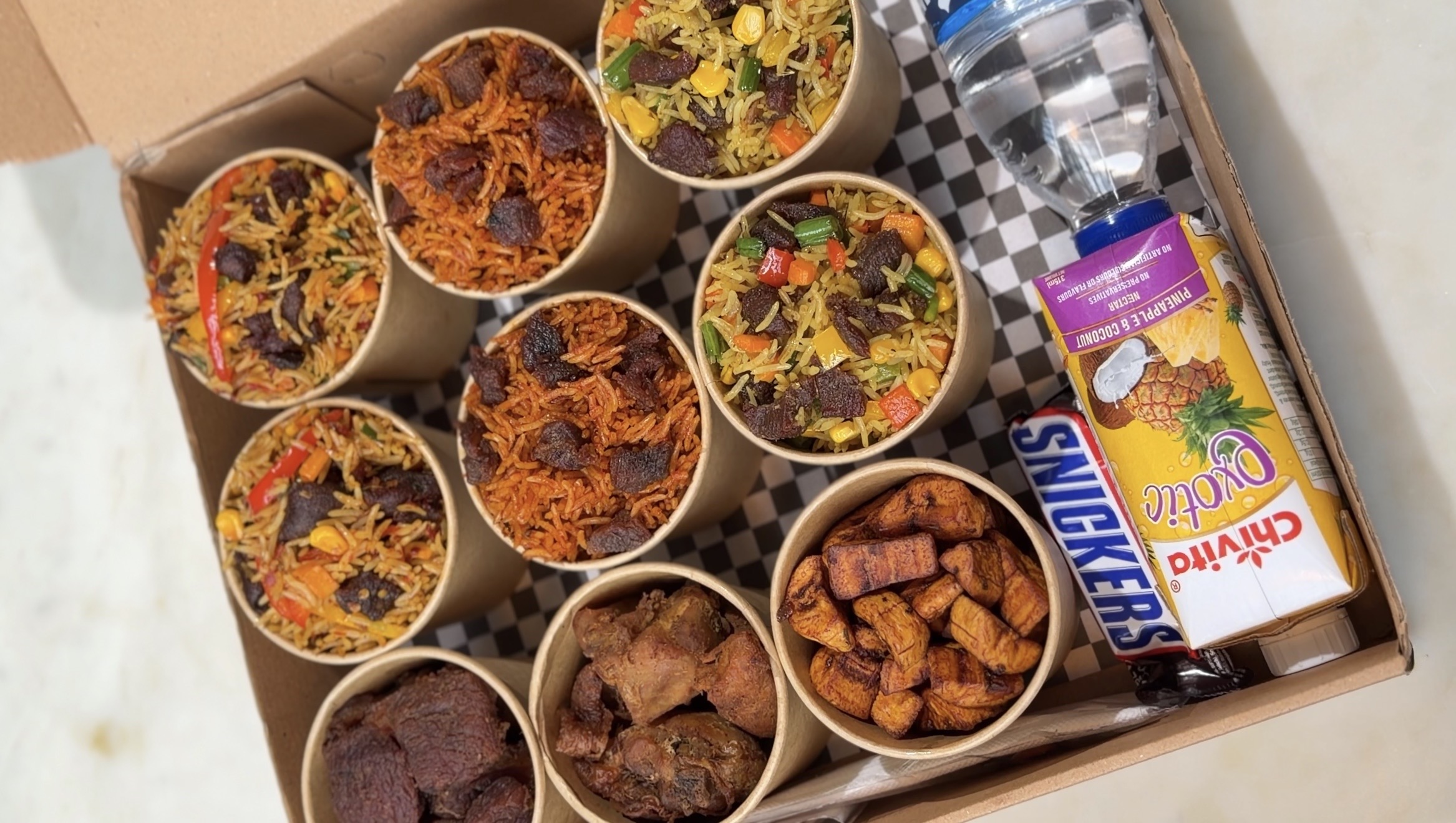 Delight Food Box opened, showing nine cups of assorted fried rice and jollof rice, grilled chicken, beef and fried plantain, plus a Snickers bar, Chivita juice and a bottle of water