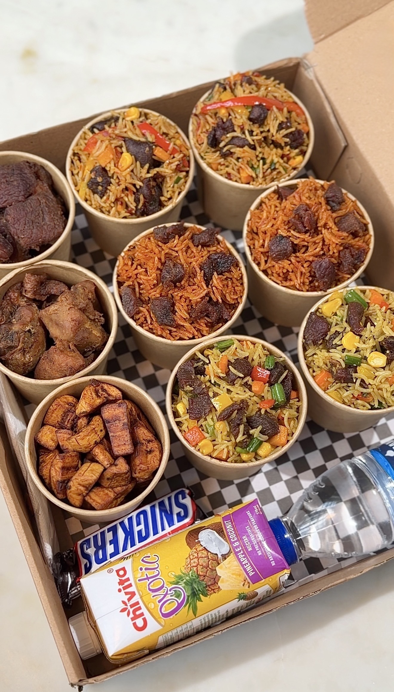 Delight Food Box opened, showing nine cups of assorted fried rice and jollof rice, grilled chicken, beef and fried plantain, plus a Snickers bar, Chivita juice and a bottle of water
